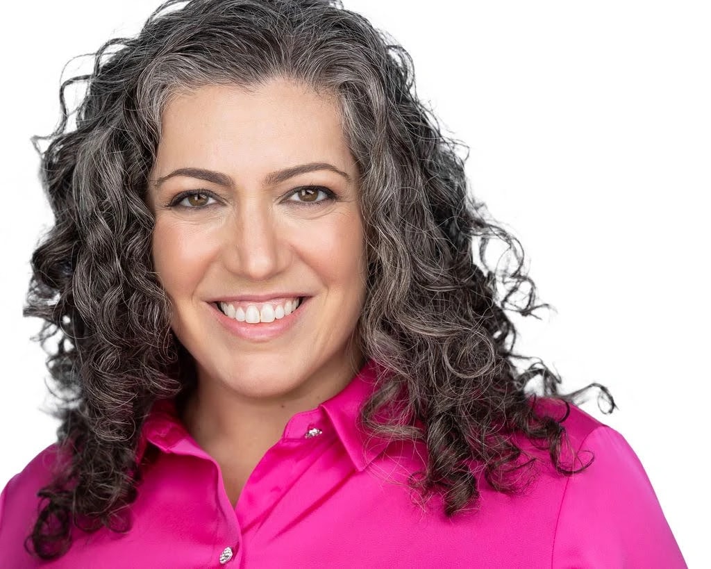 Portrait of a smiling woman with curly gray-streaked hair wearing a bright pink blouse against a white background.
