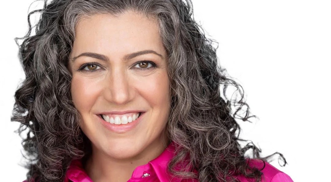 Portrait of a smiling woman with curly gray-streaked hair wearing a bright pink blouse against a white background.