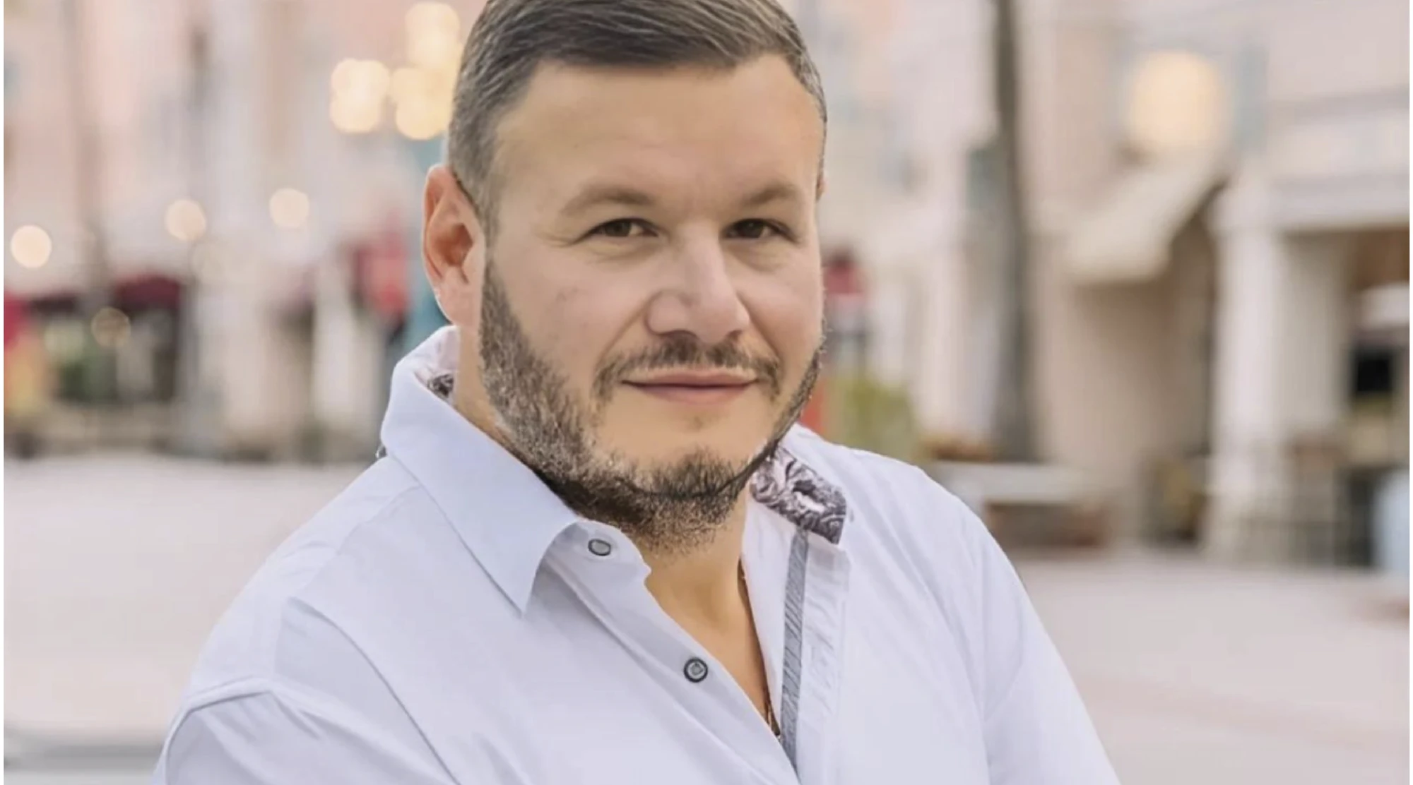 Portrait of a smiling man with a short beard wearing a light blue button-down shirt, standing on a city street at dusk.