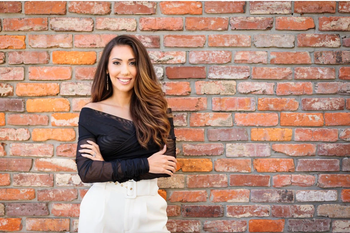 Portrait of a smiling woman with long dark hair, arms crossed, standing in front of a red brick wall.