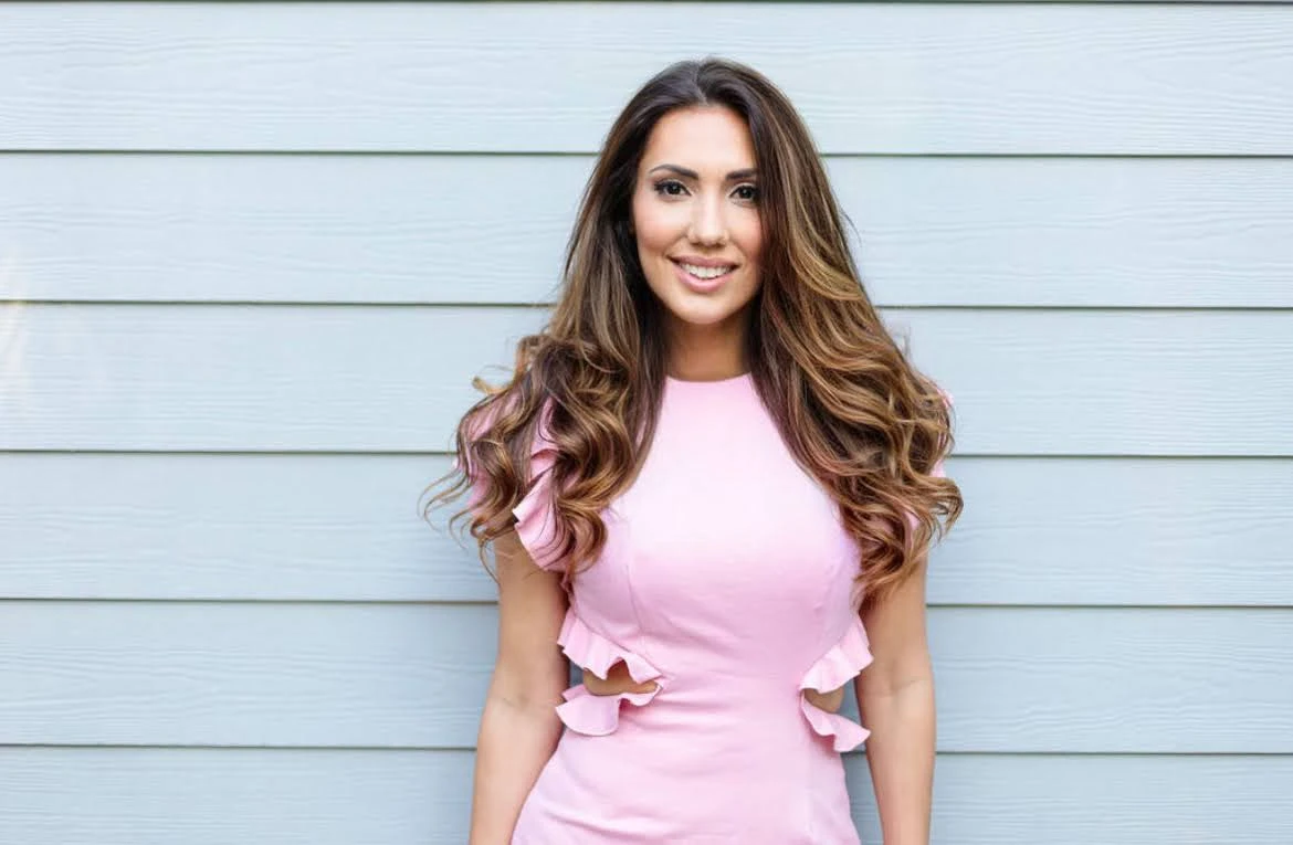 Portrait of a smiling woman with long wavy hair wearing a pink ruffled dress against a light blue paneled wall.