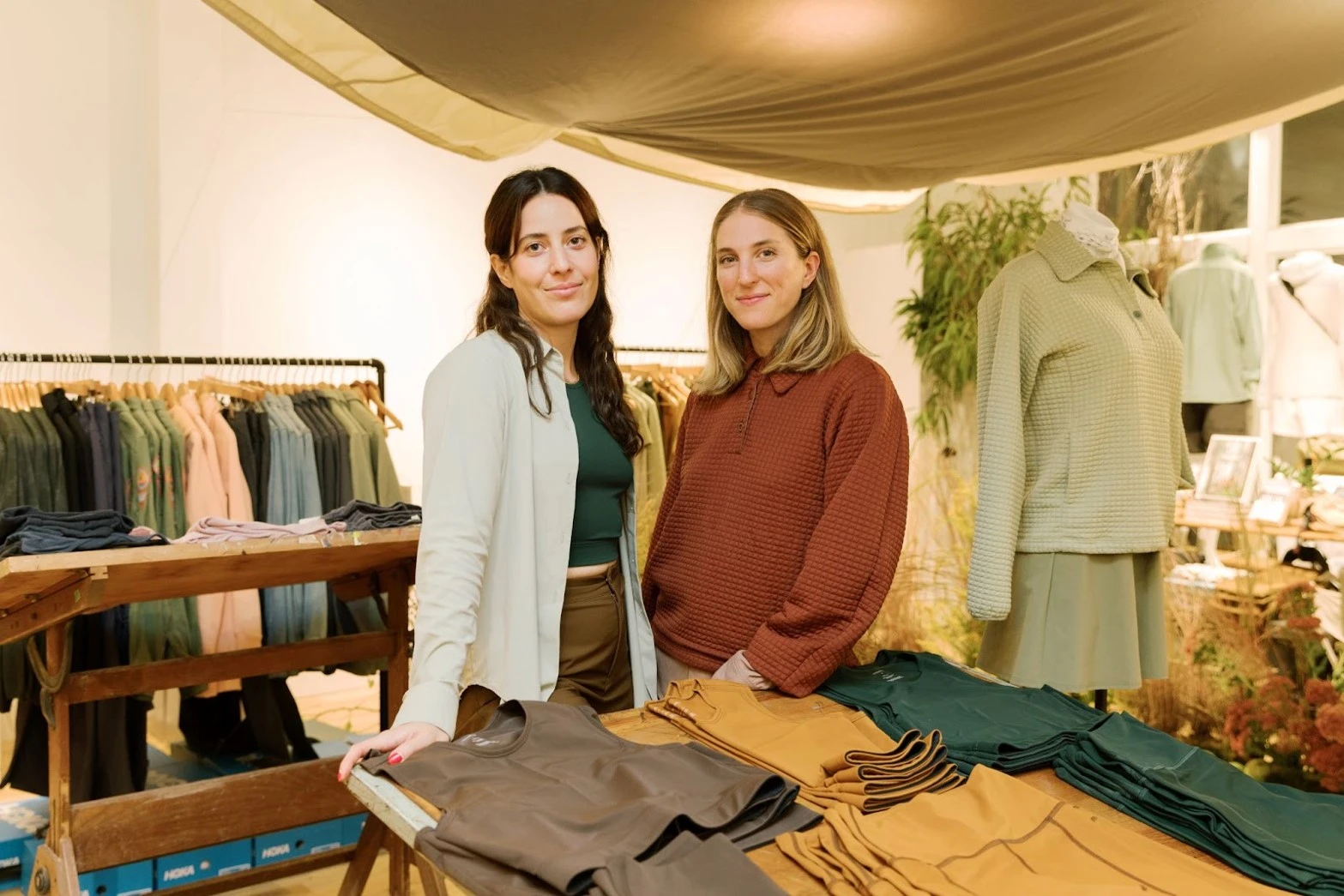 Two women standing together at a clothing display table filled with folded olive, green and mustard apparel under a canvas awning.