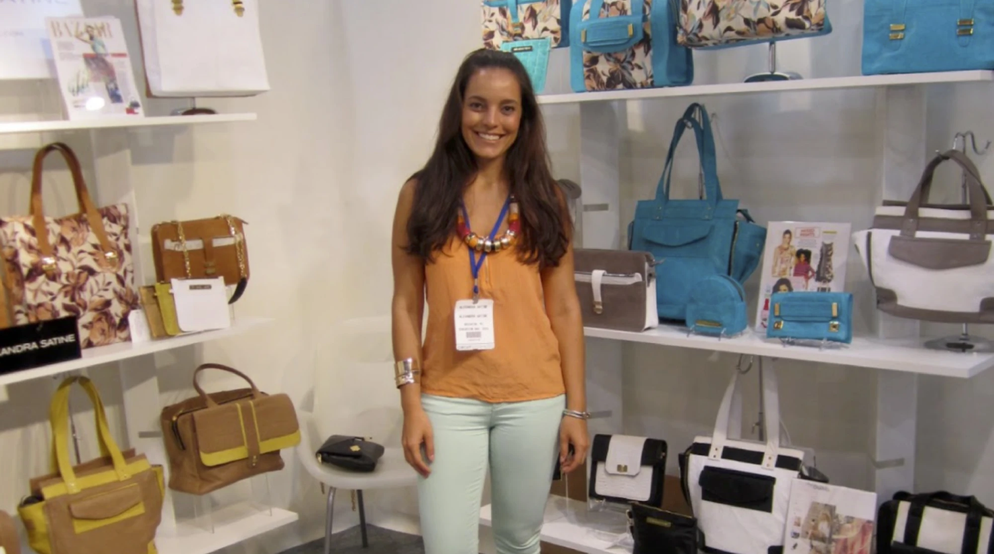 A smiling woman with a lanyard standing among shelves of colorful handbags on display at a trade show booth.