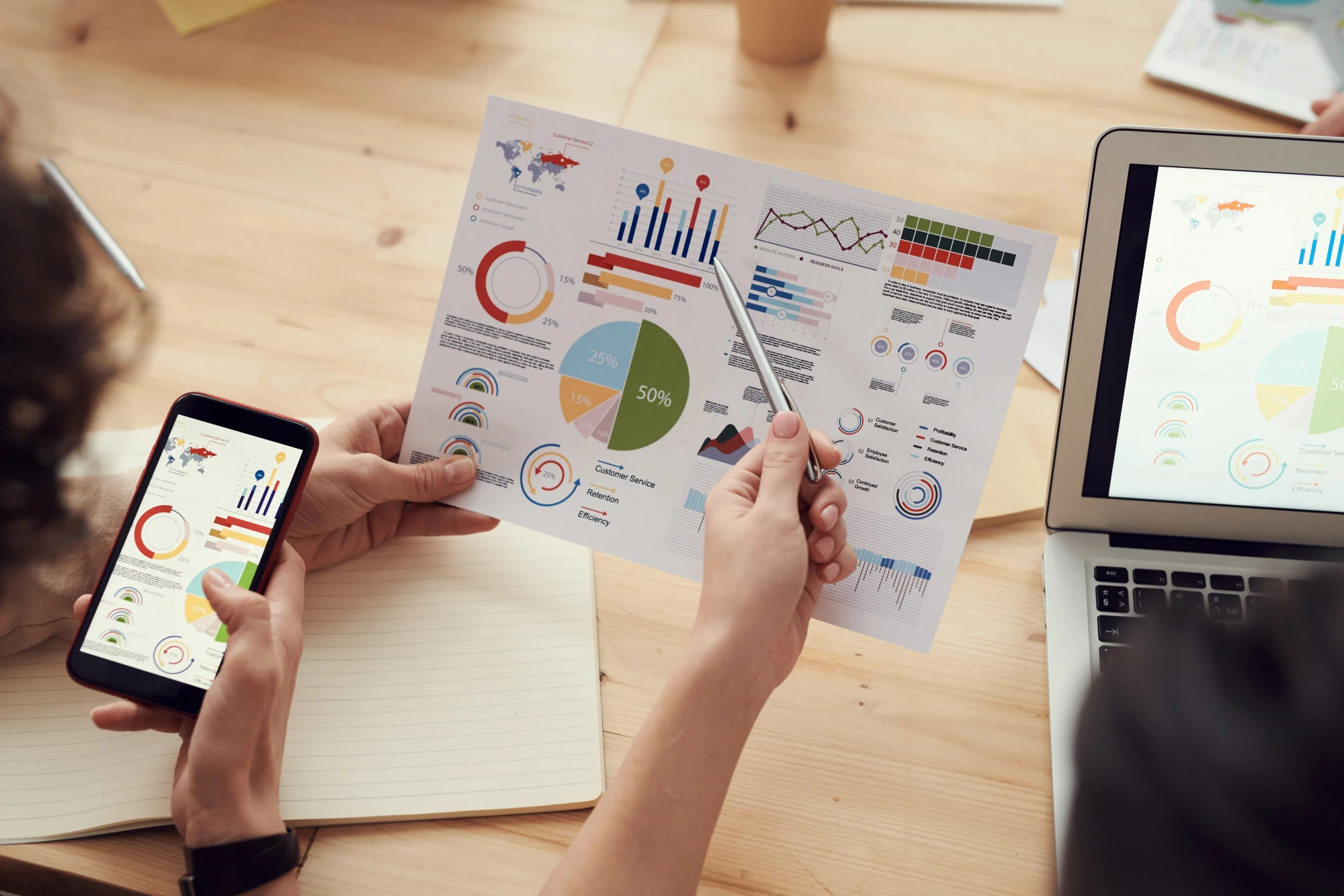 Overhead view of two people's hands holding a phone and a printed infographic with pie charts and bar graphs, beside an open laptop showing the same data.