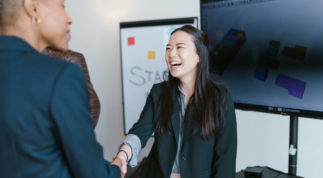 Two women shaking hands and smiling in an office, with a whiteboard and a screen showing 3D design blocks in the background.