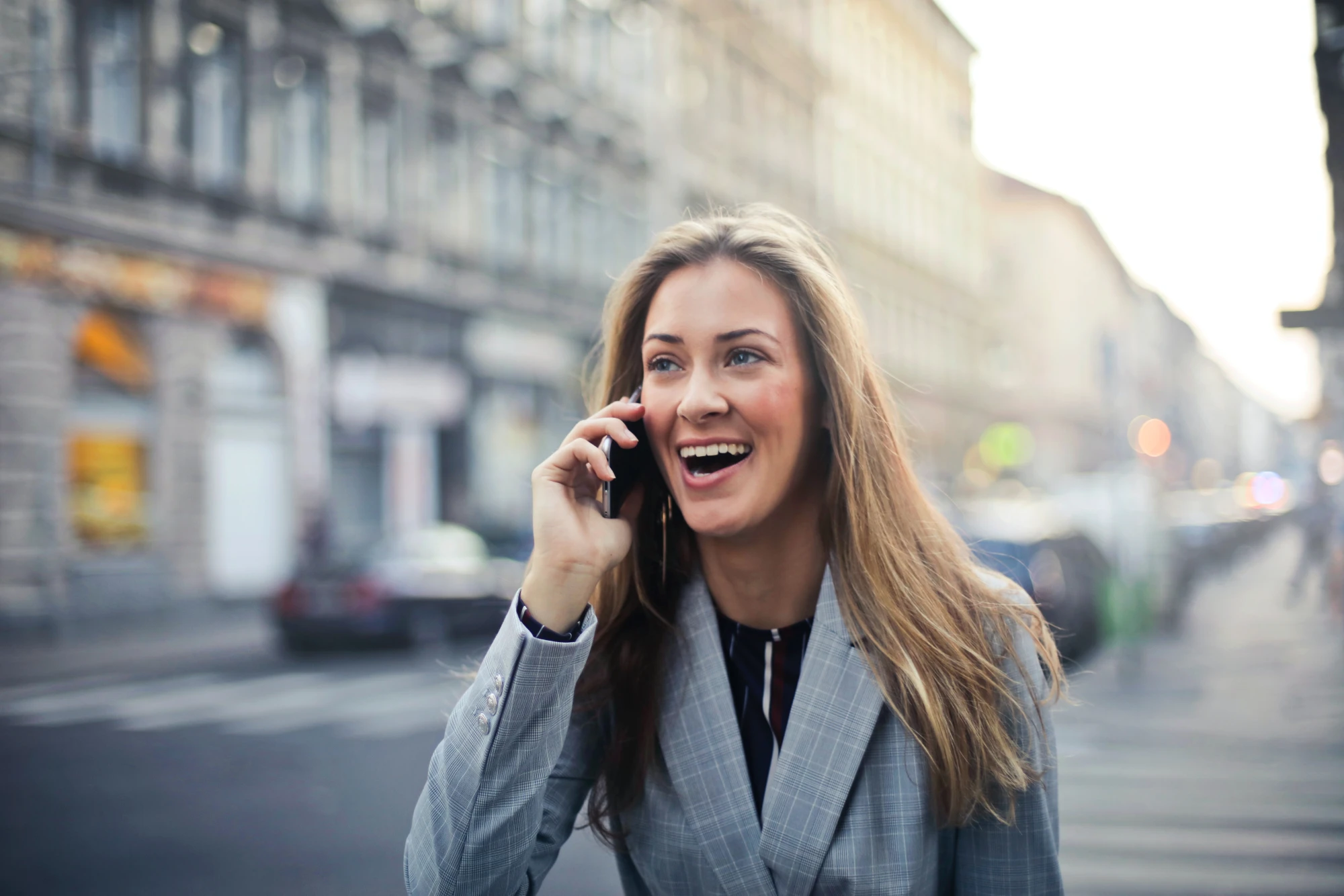 A smiling businesswoman in a plaid blazer talking on her phone while standing on a city street.