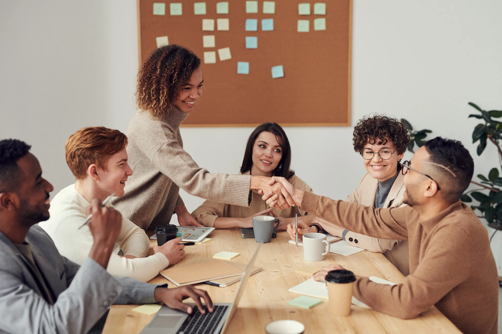 A group of colleagues around a conference table shaking hands and smiling, with a sticky-note board and a screen showing 3D design blocks behind them.
