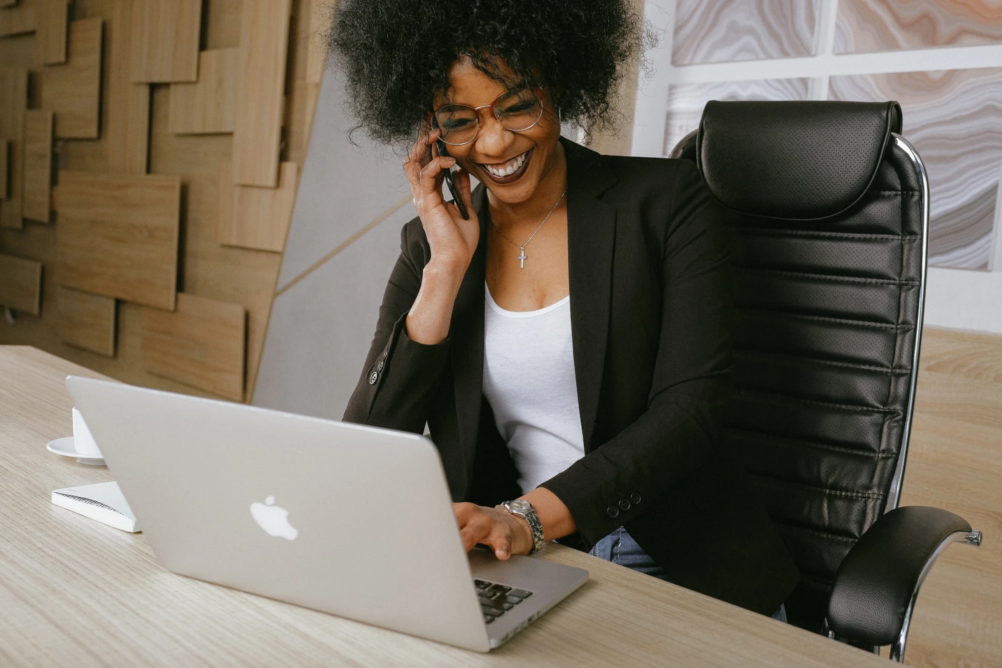 A smiling woman in a blazer talking on her phone while working on a laptop at a wooden desk in a modern office.